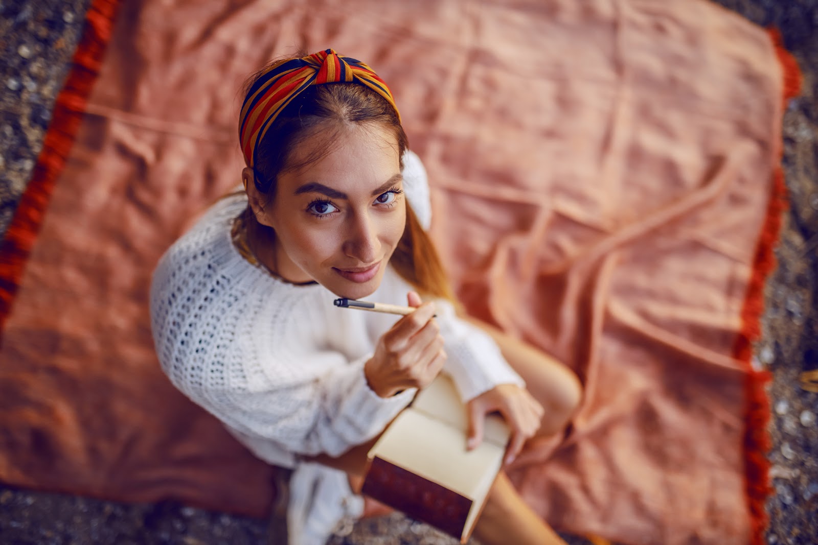 Top view of attractive caucasian brunette in sweater and with headband sitting on blanket outdoors and holding pen and diary in hands while looking at camera.