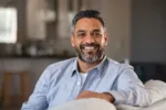 Portrait of happy mid adult man sitting on sofa at home. Handsome latin man in casual relaxing on couch and smiling. Cheerful indian guy looking at camera.