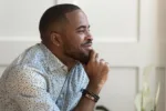 Dreamy calm african american young man looking away contemplating daydreaming sitting at home, thoughtful happy black guy dreaming thinking of good future, hoping meditating enjoy peace of mind