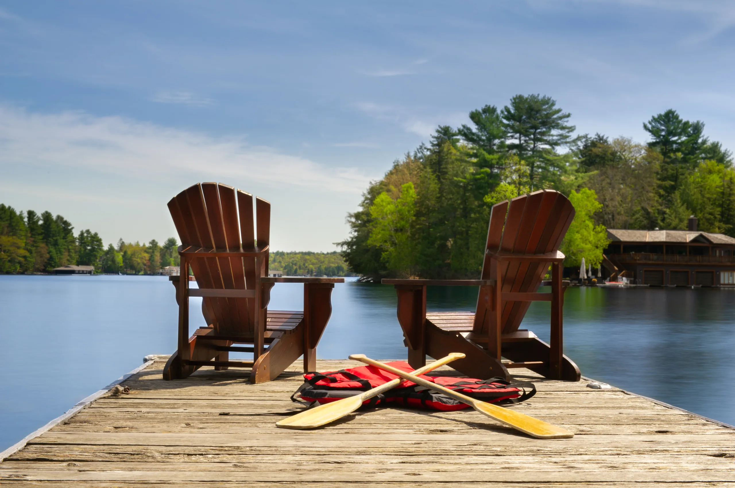 Two Adirondack chairs on a wooden dock facing the blue water of a lake in Muskoka, Ontario Canada. Canoe paddles and life jackets are on the dock. A cottage nestled between green trees is visible.