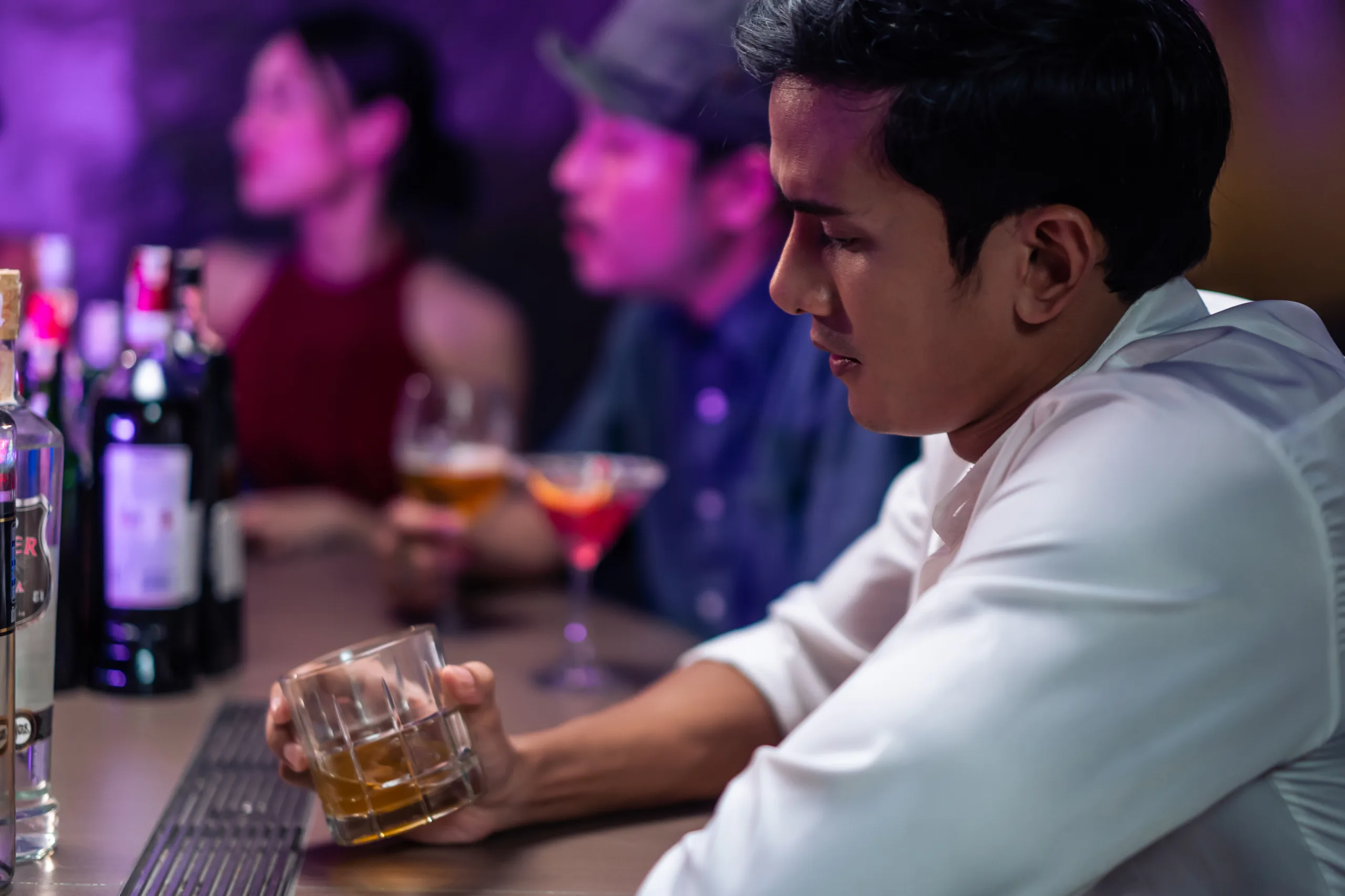 Asian depression man feeling heart broken and drinking beer in a bar. Attractive male sitting on counter bar, holding a bottle of alcohol feeling loneliness, drunk and hangover alone at night club.