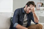 Man looking depressed while sitting alone with his head in his hand on his living room sofa at home