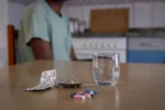 Close up of some medicines and a glass of water, person sitting behind, concept of drug consumption