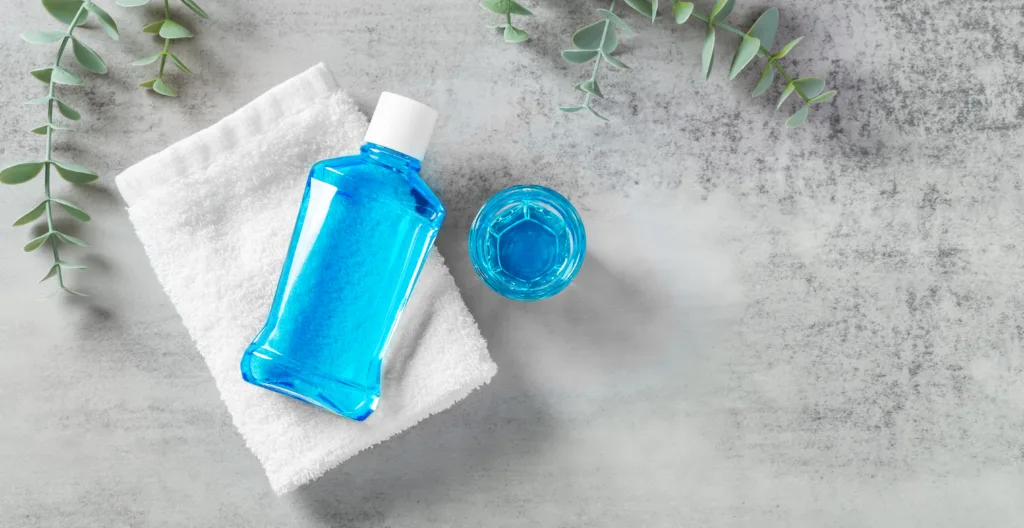 Mouthwash on white towel with cup and eucalyptus on grey table, flatlay, oral care background with copy space