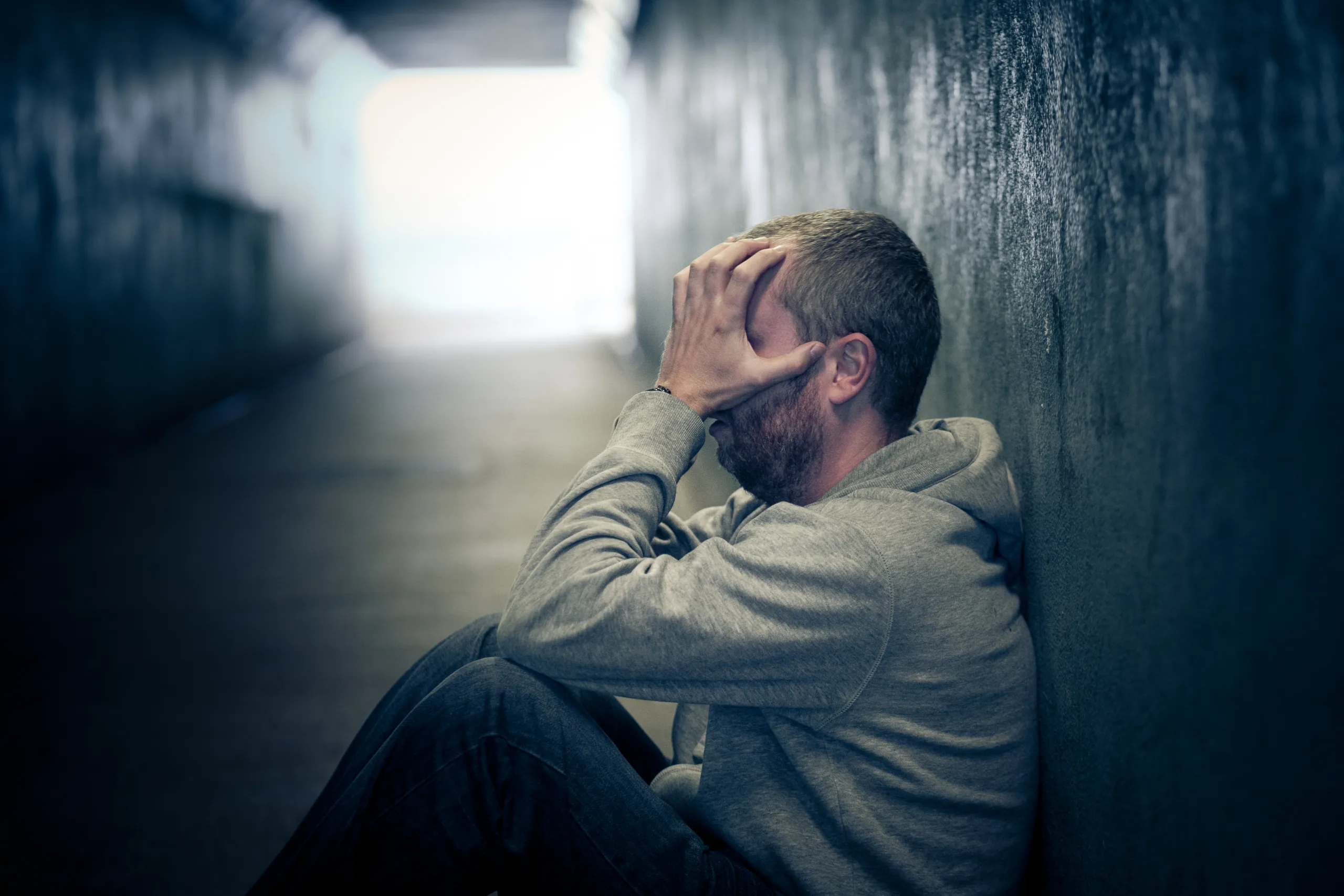 Profile view of a young homeless male - in his late 20s or early 30s - sitting in a dark, damp subway tunnel, his knees drawn up and his hands covering his face in desperation and despair. He is hungry, lonely and desperate addicted to fentanyl and xylazine. The man has short cropped hair and an unkempt beard. There is a look of sadness, loneliness and desperation in his eyes. He is sitting on a piece of old cardboard Horizontal image with room for copy space.
