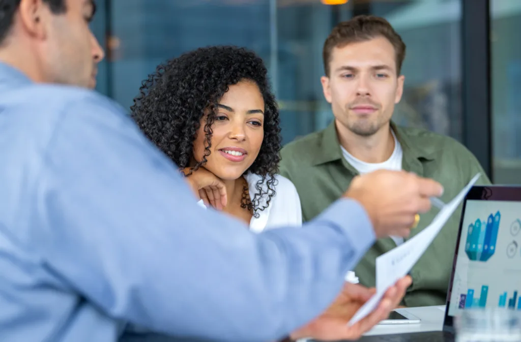 Couple listening to a businessman, banker or financial advisor who has a laptop computer showing financial charts and graphs. He is showing them paperwork contract or information.