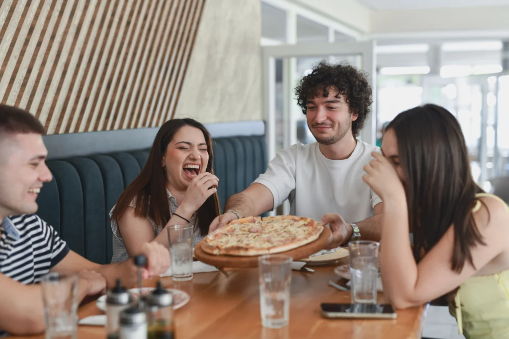 Smiling Male Friend Sharing Hot Pizza With Friends While Female Friends Are Laughing Loudly